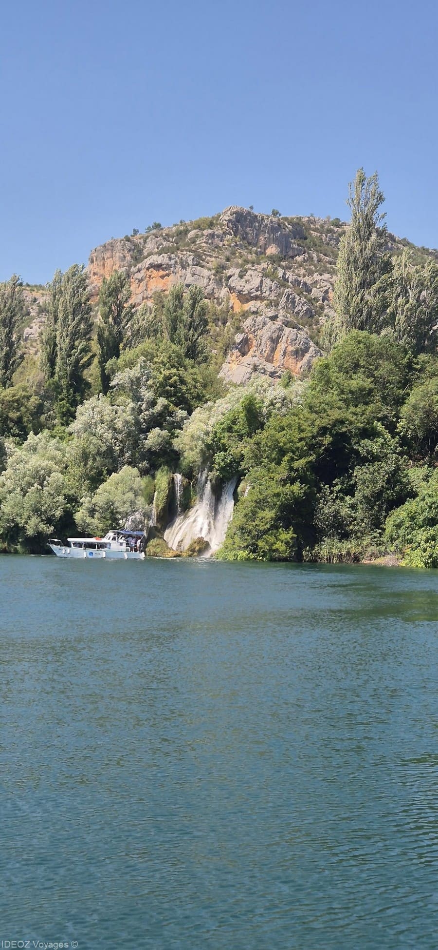 Bateau sur la rivière Krka devant une cascade bordée d’arbres et de falaises