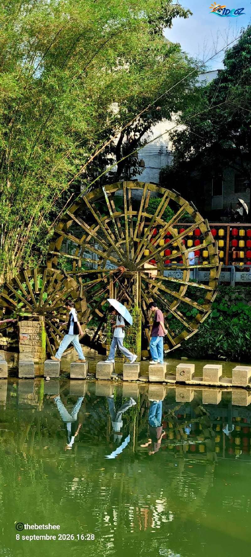 Trois personnes traversent un cours d'eau sur des plots de pierre, devant deux grandes roues à eau en bois dans le village ancien de Huangyao.